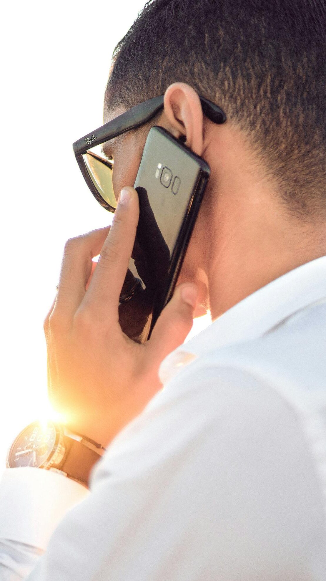 Man outdoors in Morocco talking on smartphone, wearing sunglasses and white shirt, backlit by sunset.